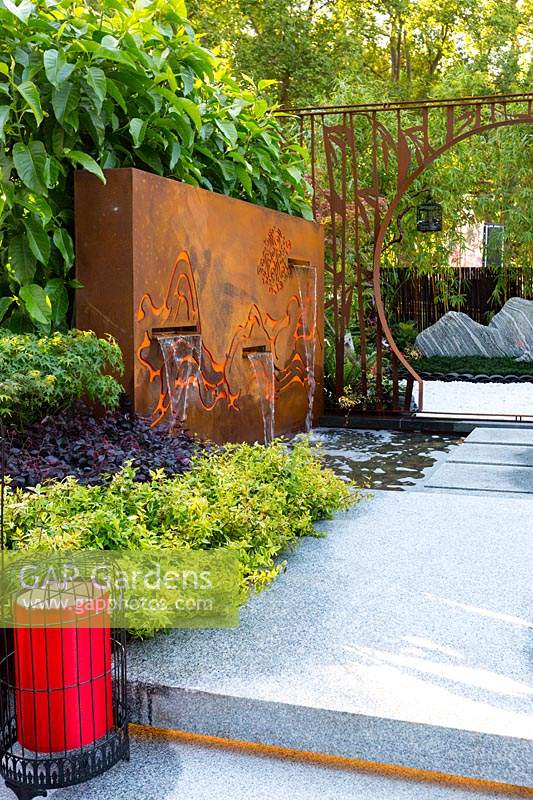 Pavilion style Chinese garden, with a freestanding water feature, moongate, stepping stones, striped paving , planter boxes, stone mountain range, printed glass panels, steps and red lanterns.
