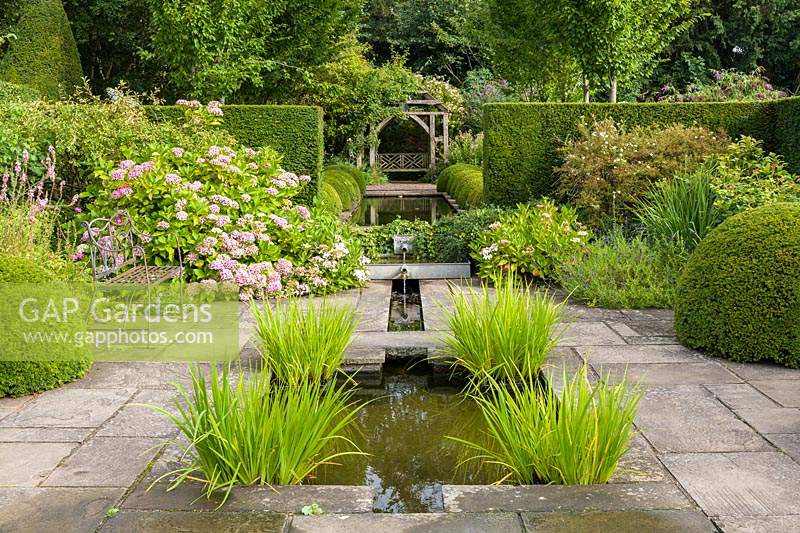 Water flows through the paved Lower Rill Garden at Wollerton Old Hall Garden, near Market Drayton, Shropshire, UK - beyond clipped box balls line the rill, with an oak gazebo in the distance