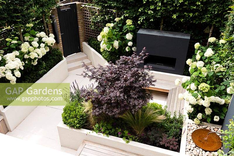 View from above of small contemporary private urban garden with porcelain paving, raised beds, outdoor fireplace, floating bench seating and water feature.