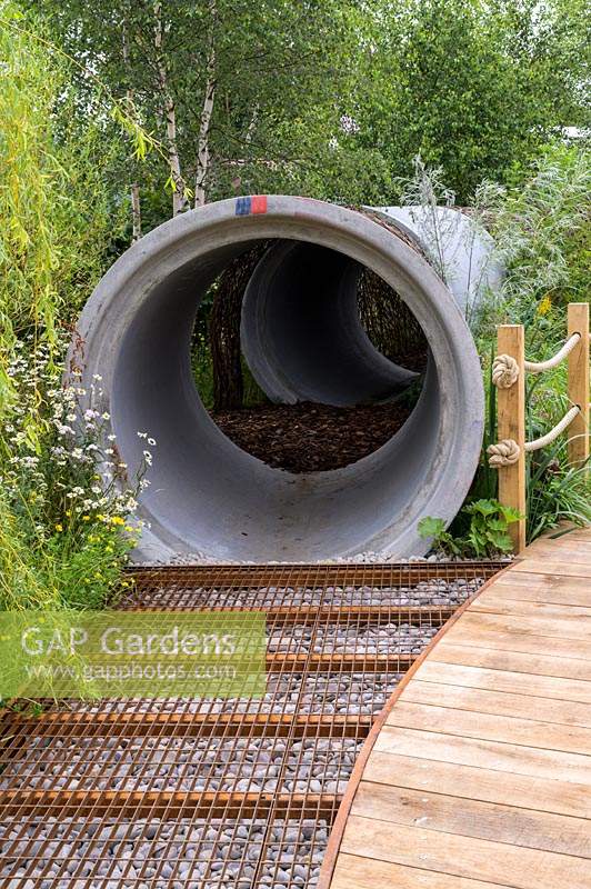 A woven willow arch covering a large water pipe with metal grid walkway filled with pebbles - The Thames Water Flourishing Future Garden - RHS Hampton Court Palace Garden Festival 2019.