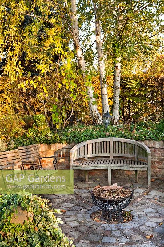 Sunken patio with circular paving around a fire pit, seating in front of a raised bed with trees and perennials