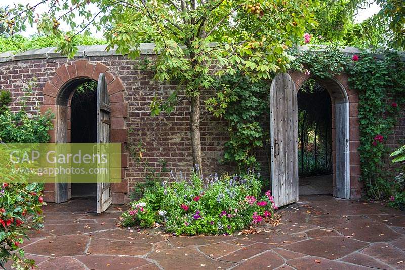 Brick walls with stone arches with wooden gates. Wall has Rosa - Climbing Rose over entrance and Aesculus glabra - Ohio Buckeye - underplanted between the gates