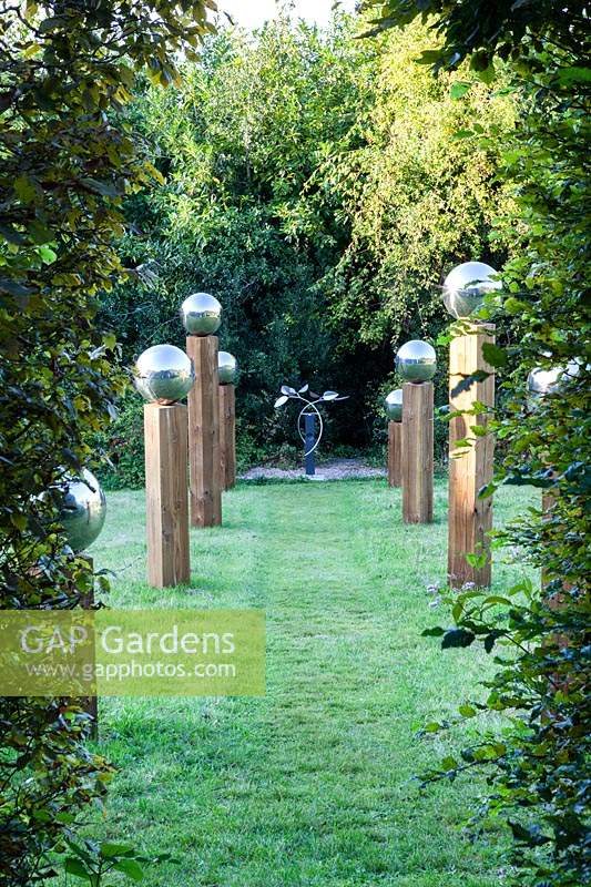 Avenue in The Meadow. Stainless steel mirror globes on top of wooden pillars of irregular height. Mobiles sculpture at the focal point is 'Octo' by Stuart Stockwell. Veddw House Garden, Monmouthshire, Wales, UK. 