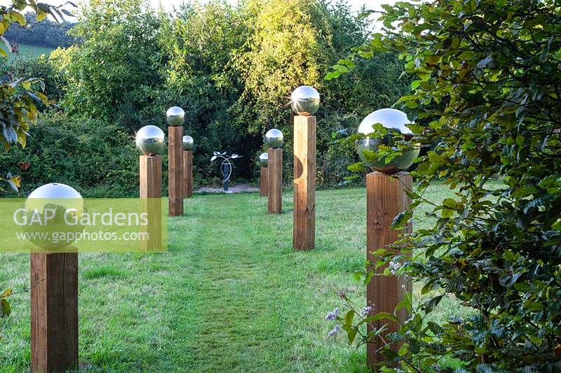 Avenue in The Meadow. Stainless steel mirror globes on top of wooden pillars of irregular height. Mobiles culpture at the focal point is 'Octo' by Stuart Stockwell.Veddw House Garden, Monmouthshire, Wales, UK. 