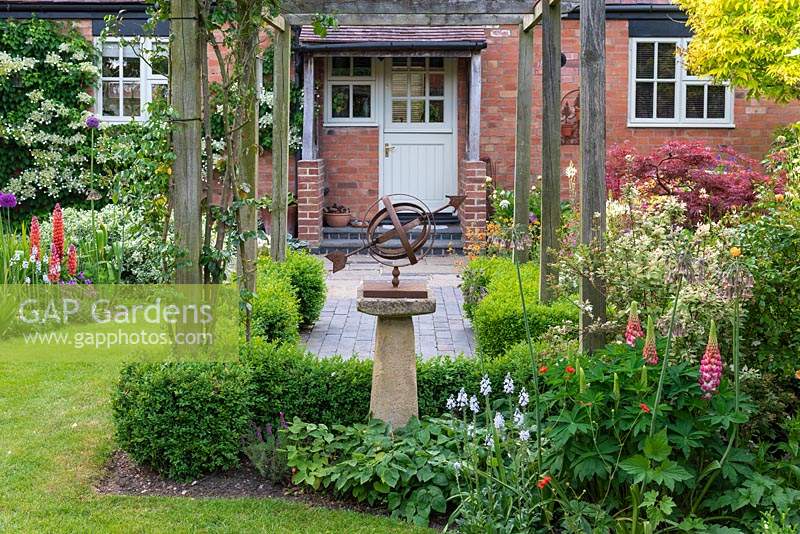 A rusted metal sundial on a stone plinth provides a focal point at the end of a brick path with wooden pergola. 