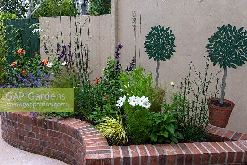 Curving, brick edged, raised bed planted with cosmos, roses, salvias, catmint and Verbena bonariensis. Behind are murals of potted bay trees on wall.