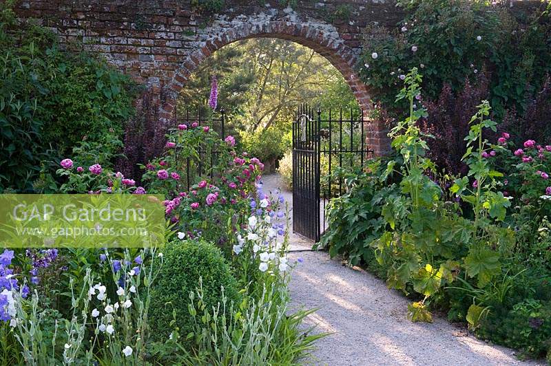 Gravel pathway through gate set in arch of old brick boundary wall by flowering summer borders. 