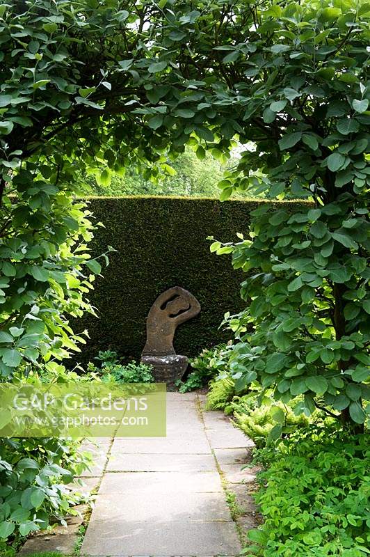 Path through arch in hedge framing sculptural stone seat by Taxus hedge 