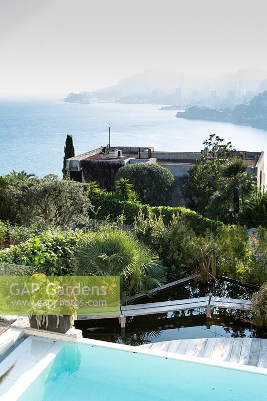View over swimming pool, garden with mix of plants including palms and outwards to the sea and coastline 