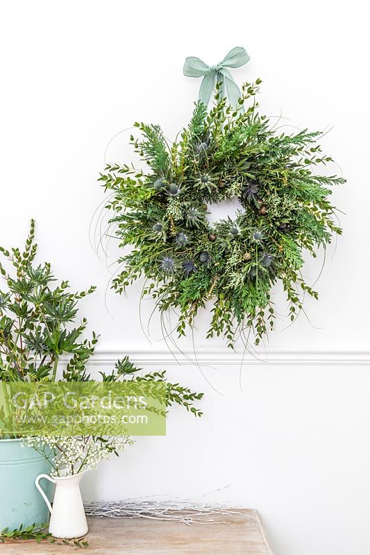 Green-themed Winter wreath embellished with blue Eryngium flowers, hung on white wall 