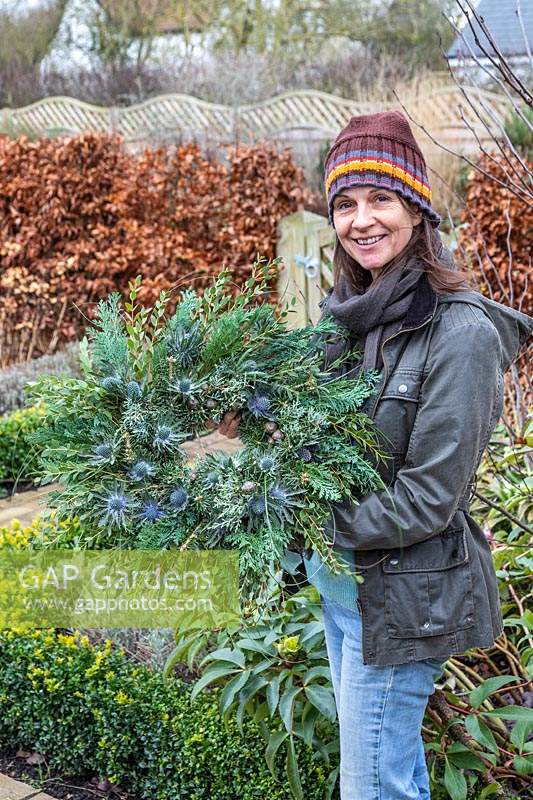 Woman holding green-themed winter wreath in garden setting. 