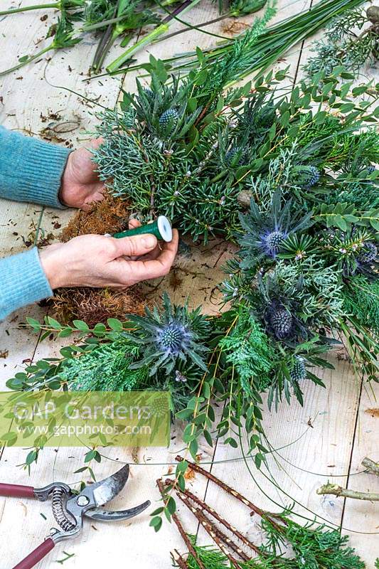 Woman attaching bundle of foliage and flowers to winter wreath with floristry wire. 