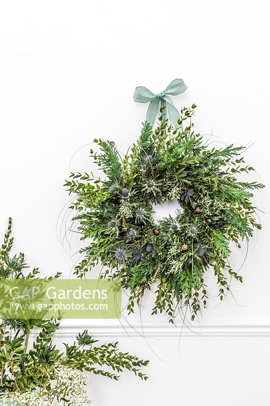 Green-themed Winter wreath embellished with blue Eryngium flowers, hung on white wall.