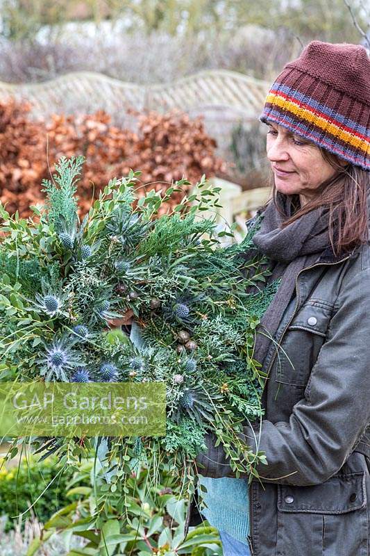 Woman holding green-themed winter wreath in garden setting.