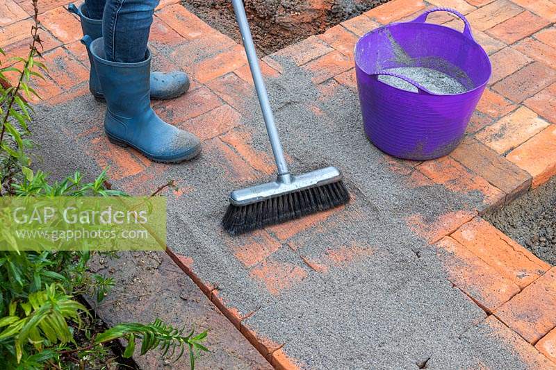 Woman using soft brush to push polymer sand into joins of newly laid brick path