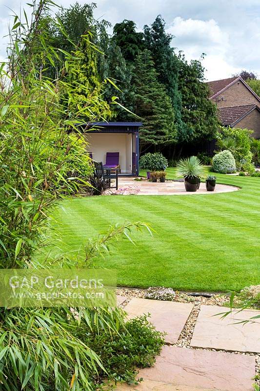 Overview of a garden, screened from neighbours by conifers, neat striped lawn, patio with pots and a summerhouse 