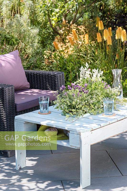 Wooden pallet table with integral sunken planter with summer planting, on slate patio in summer, accompanied by outdoor chairs.