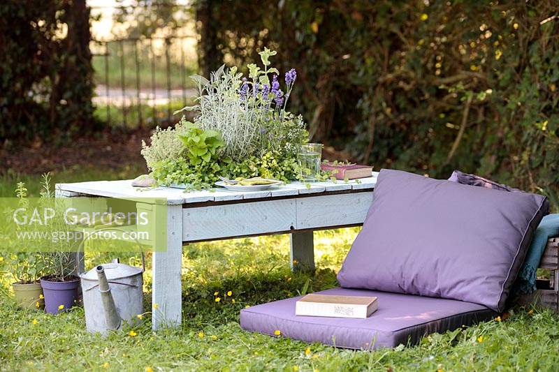 Table made out of a wooden pallet with central planter planted with mixed herbs in summer country garden with cushions.
