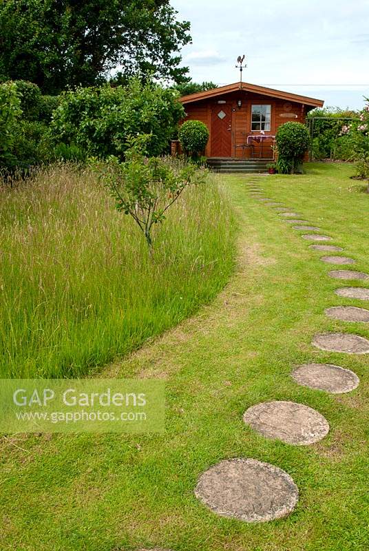 Tall grass around fruit trees left for the benefit of wildlife and insects, with path of stepping stones leading to distant summerhouse - Open Gardens Day, Kelsale, Suffolk