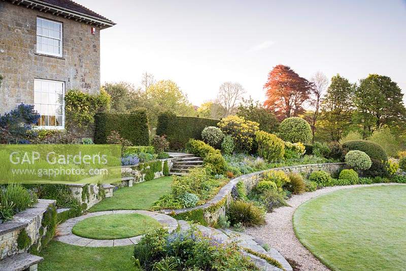 A view from the upper terrace across curving walls, lawns and borders towards clipped shrubs and lawn below.