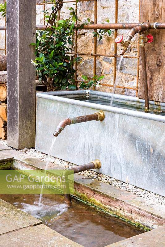 Galvanised drinking trough, copper pipes and taps used as a water feature below a timber pergola in the courtyard garden at Am Brook Meadow, Devon in August. 