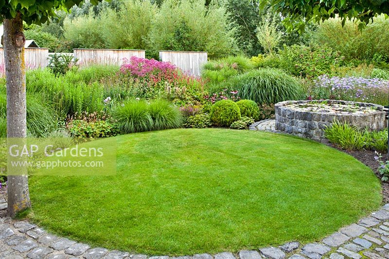 Small circular lawn with circular pond and perennial border beyond, edged with setts