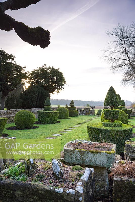 The Topiary Garden edged with stone troughs, a wall and urns, with countryside beyond at Rodmarton Manor, Glos, UK. 