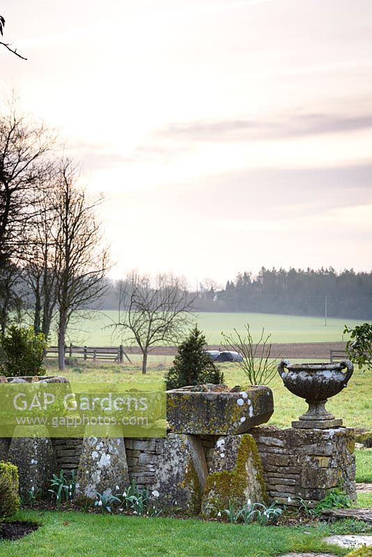 Low stone wall displayed with troughs and classic urns, with view to countryside beyond at Rodmarton Manor, Glos, UK. 