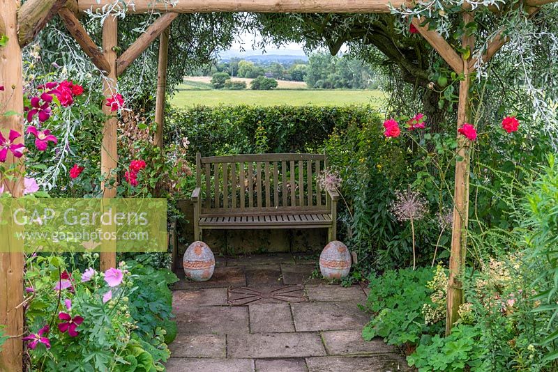 View along a path beneath a rustic pergola to a bench, with views over Blackmore Vale beyond.

