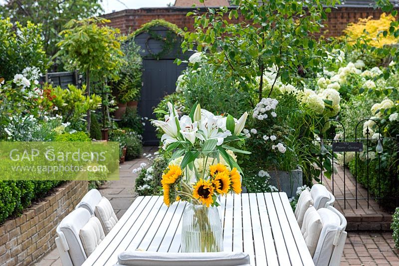 Dining table with vases of sunflowers and white lilies of sunflowers and white lilies against backdrop of white agapanthus, fuchsia,  hydrangeas, geraniums, cosmos, roses, gaura, busy lizzies and silver birches.