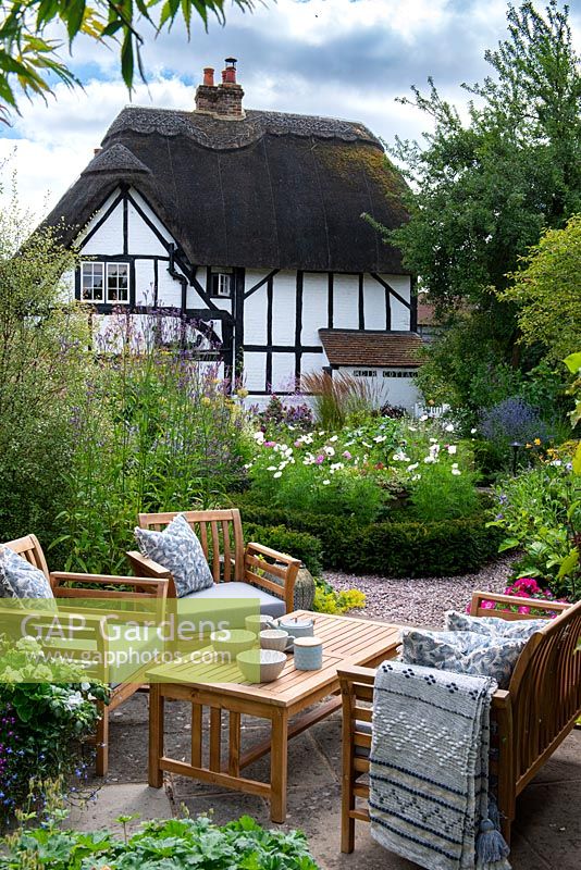 View over seating area to yew-edged beds of tall perennials, ornamental grasses and Cosmos, thatched cottage beyond