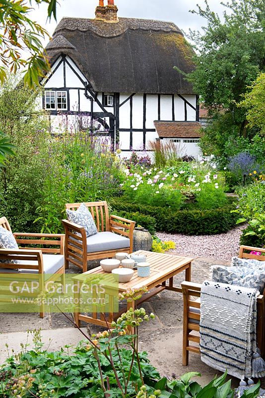 View over a seating area to yew-edged beds of tall perennials, ornamental grasses and Cosmos, thatched cottage beyond
