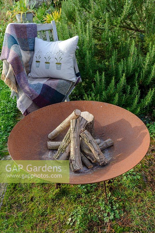 A Fire bowl made from rusted corten steel, a wooden garden chair with a wool rug and a cushion with a snowdrop design by a large rosemary bush.
