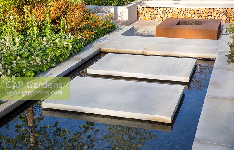 Large sawn Yorkstone floating stepping stone slabs over a water feature leading to a sunken garden with corten steel fire pit. The Sunken Retreat, RHS Malvern Spring Festival, 2016. Design: Ann Walker for Graduate Gardeners 