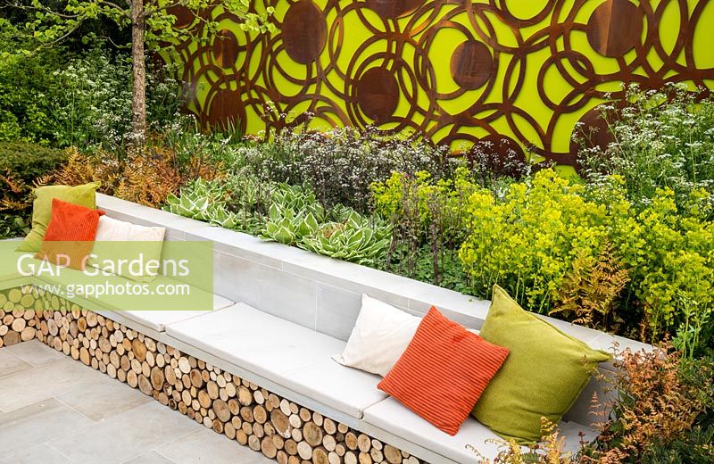 Large sawn Yorkstone stone bench with colourful cushions and log storage underneath. The Sunken Retreat. RHS Malvern Spring Festival, 2016. Design: Ann Walker for Graduate Gardeners
