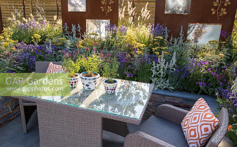 Outdoor acrylic rattan dining furniture with table and chairs with orange cushions on patio surrounded by a lucent copper slate raised bed with colourful planting of yellow and purple flowering plants, ornamental grasses with rusted corten steel screens. The RNIB Community Garden. RHS Hampton Court Flower Show July 2018  - Designer: Steve Dimmock and Paula Holland - Sponsor RNIB, Magus Private Wealth 