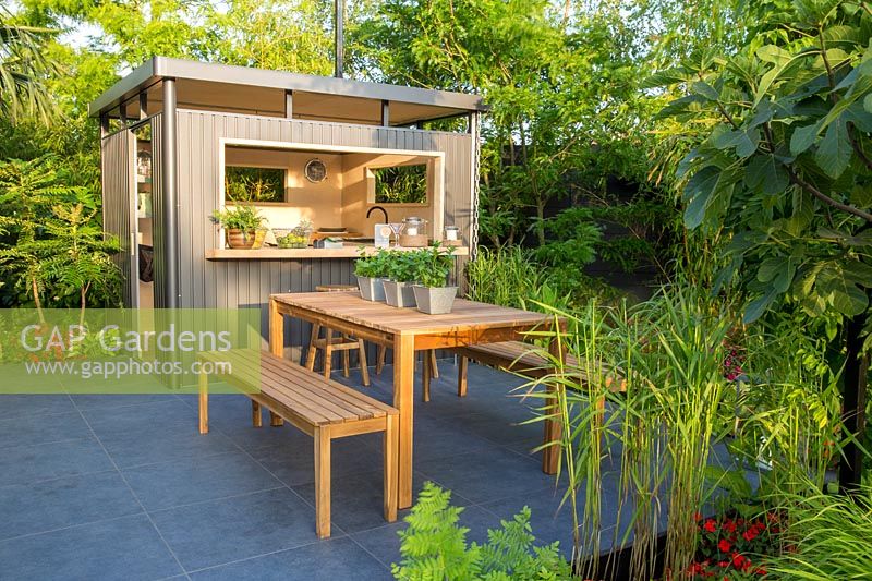 An outdoor entertaining space with metal outdoor kitchen bar set on porcelain stone patio with wooden bench and table. B and Q Bursting Busy Lizzie Garden