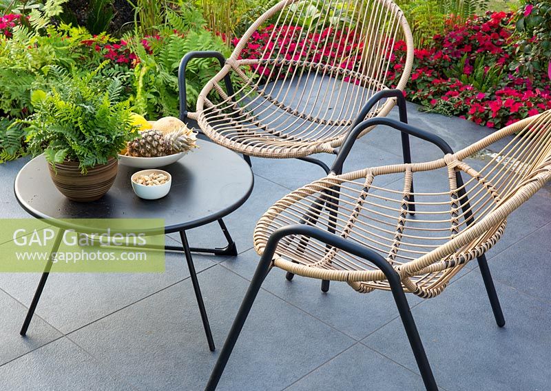 Metal framed cane chairs and black metal table on a porcelain stone patio with Impatiens 'Imara' in borders with ferns. B and Q's Bursting Busy Lizzie Garden
