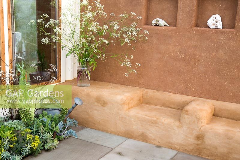 Cob and clay plastered wall with shelves and seating area small patio - border planted with herbs, kale and a glass vase with cow parsley. An Artist's Studio Home - Green Living Spaces. RHS Malvern Spring Festival May 2019 -  Design: Jessica Makins 