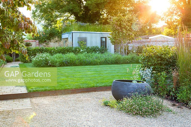 A sunlit contemporary city garden with gravelledpatio, sculptural pond, raised lawn, wildflower border and green-roofed garden room.