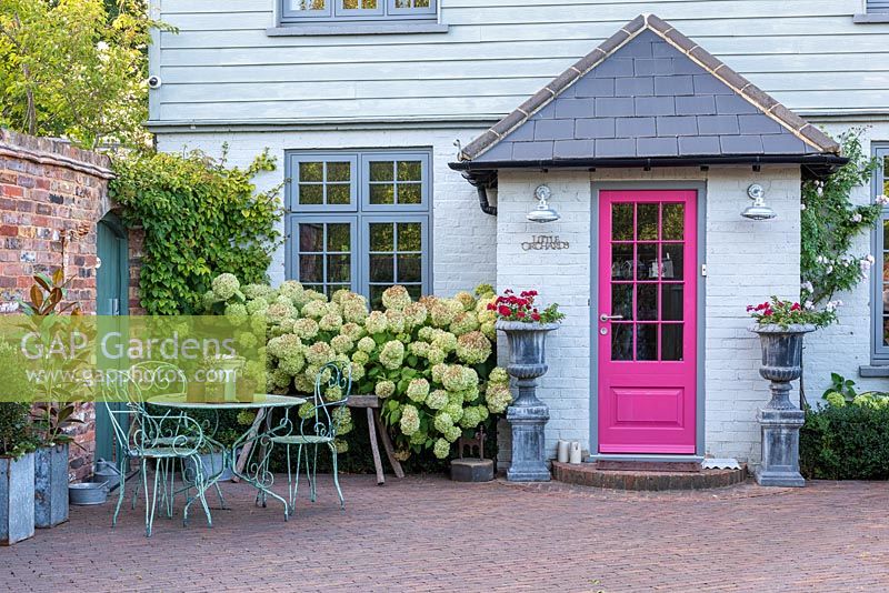 A contemporary country cottage front garden planted with Hydrangea 'Limelight, patio seating area and urns with annual geraniums.