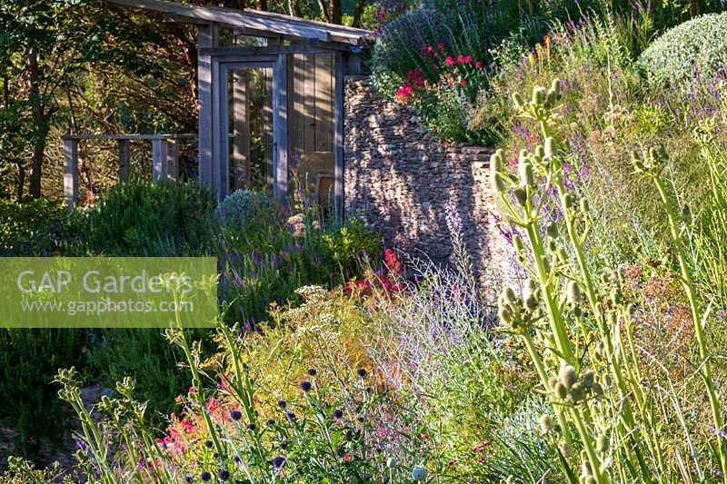 Garden on slope with flower bed in sun and shaded stone wall terrace and summerhouse
