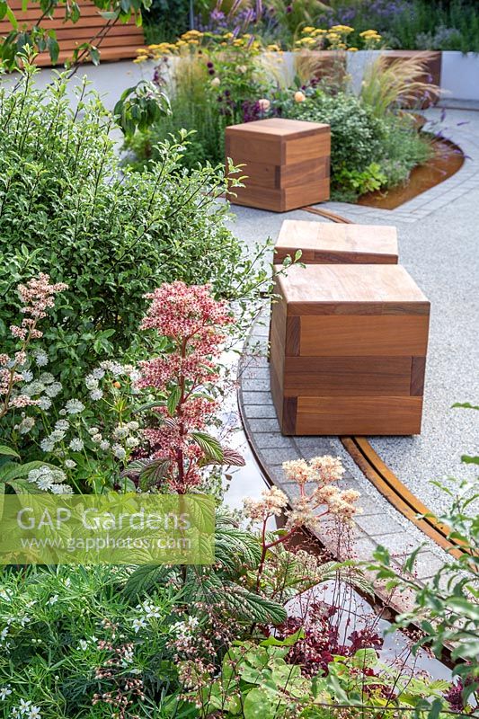 View through public space with moveable wooden cube seating on a rail, mixed summer borders edged with rill water feature. The Crest Nicholson Livewell Garden - Hampton Court Flower Show 2019 