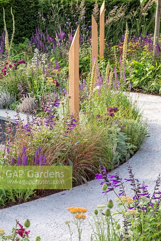 Compacted path spiralling around beds full of colourful summer perennials with inscribed wooden posts. The Cancer Research UK Pledge Pathway to Progress - Hampton Court Flower Festival, 2019.