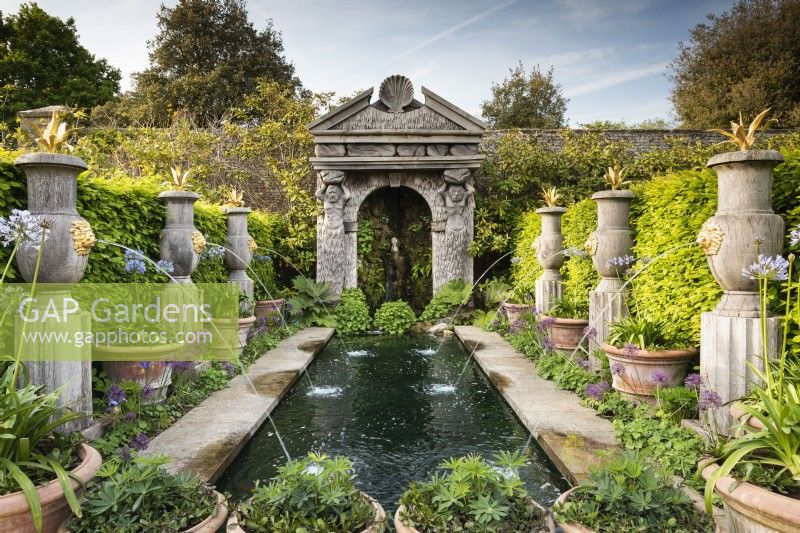 The Collector Earl's Garden at Arundel Castle, West Sussex in May. Designed by Isabel and Julian Bannermann. Green oak urns, pillars and a grotto guarded by sea gods frame water representing the alegorical River Arun. Gilded lions and agaves adorn the urns, and planting includes alliums, agapanthus and Alchemilla mollis.