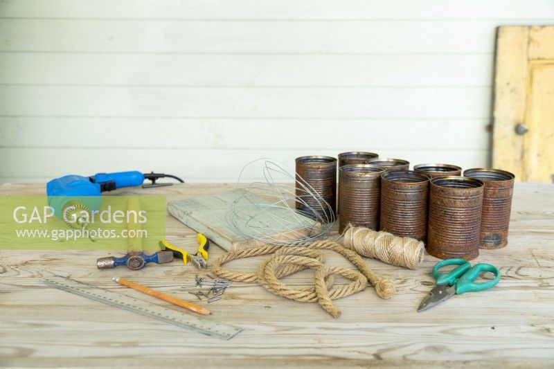 Drill, hammer, nails, pliers, scissors, pencil, ruler, wooden board, garden wire, sandpaper, rope, string and tin cans laid out on a wooden surface