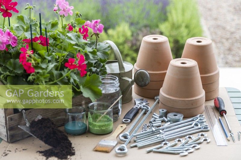 Steel rods, tensioners, hooks, connectors, nuts, washers, screwdriver, screws, spanner, watering can, pencil, terracotta pots, paint, brushes, plants and compost laid out on a table