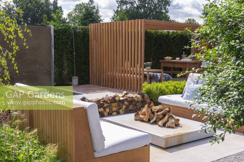 The Viking Friluftsliv Garden. View across a seating area with sofas to an outdoor dining and kitchen area, beneath an iroko pergola.