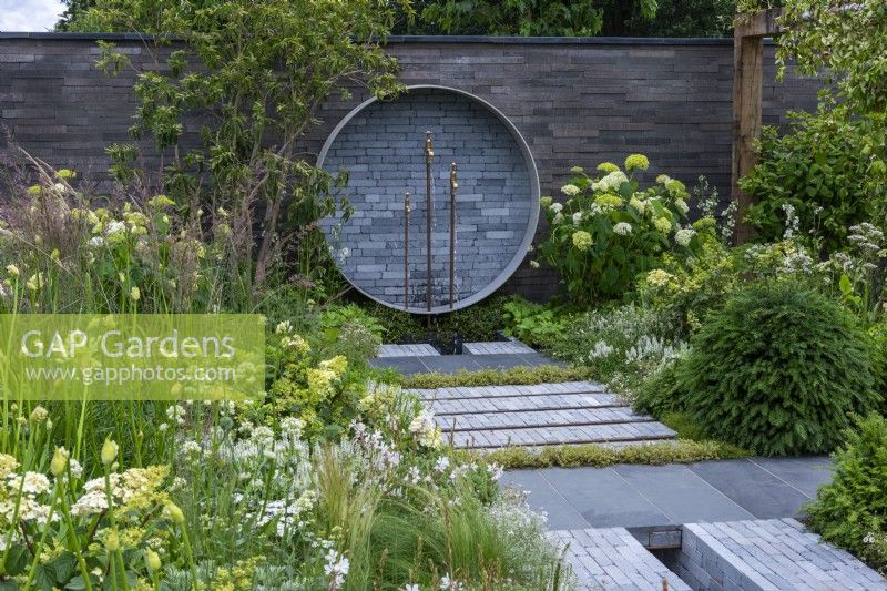 A Place to Meet Again. Hampton Court Flower Festival 2021. An urban courtyard is planted in restful tones of grey, green and white. A circular water feature on the wall upcycles pipes and taps.