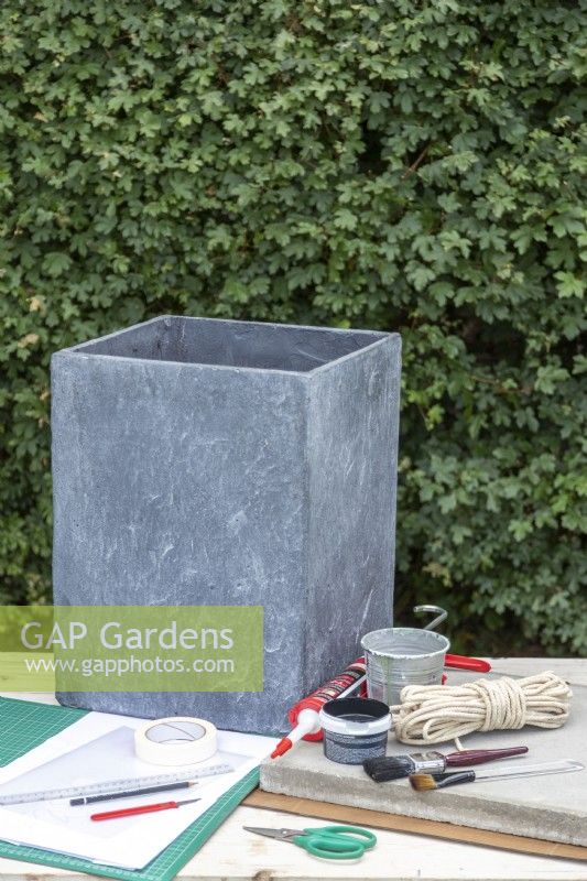 Large stone plant pot, stone slab, sash cord, masonry paint, sealant gun, adhesive, two paint brushes, craft knife, pencil, ruler, paper, masking tape, clear plastic sheet and a cutting mat laid out on a table